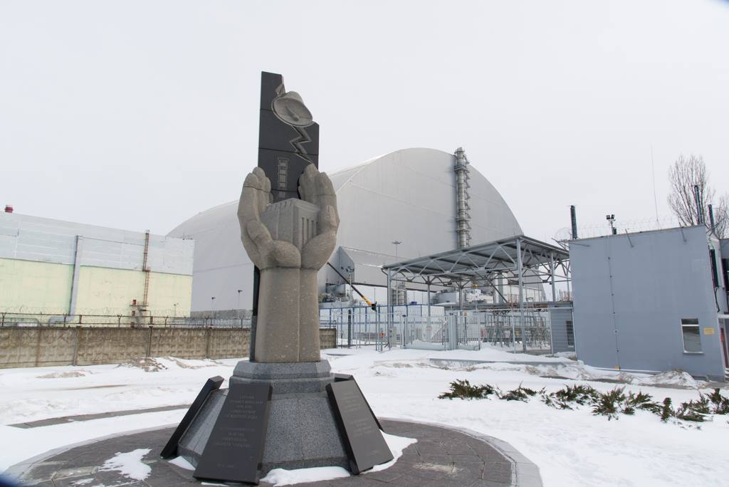 Photo of the memorial monument of two hands holding a structure. The monument sits in front of the now sealed sarcophagus which covers Reactor 4