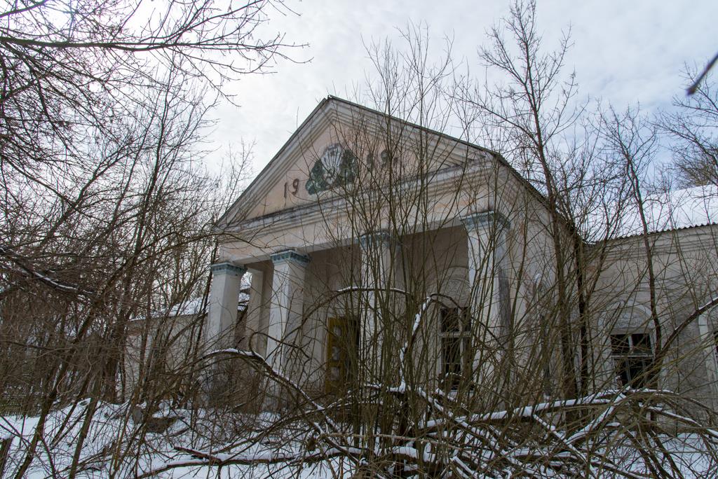 Photo of the Palace of Culture in a small village within the 30 km Chernobyl Exclusion zone. An imposing building with large white large pillars, still standing strong amongst the overgrown trees. 