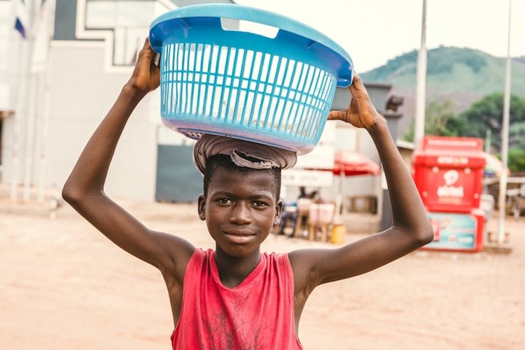 Photo of a young boy holding a washing basket it on his head as he goes about his daily routine