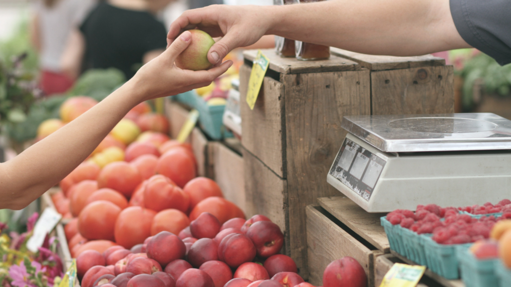 A vendor handing an apple to a shopper in a farmers market