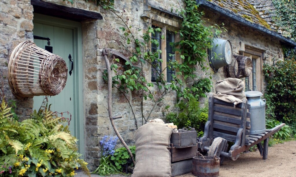 An old Cotswold cottage complete with a wicker basket, an old style wheelbarrow and a milk carrier