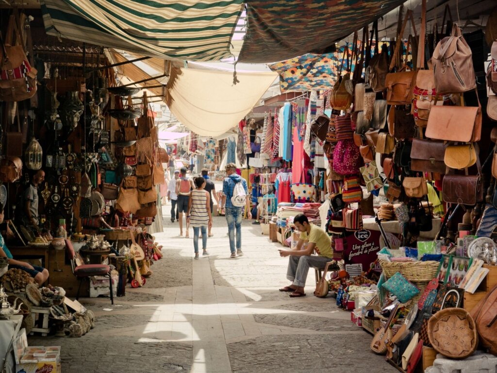 A souk in the shadows as the sun tries to penetrate between the gaps in the cloth verandas. A man is sitting on a stool looking at his phone as shoppers walk away down the corridor amongst the many wears on sale. 