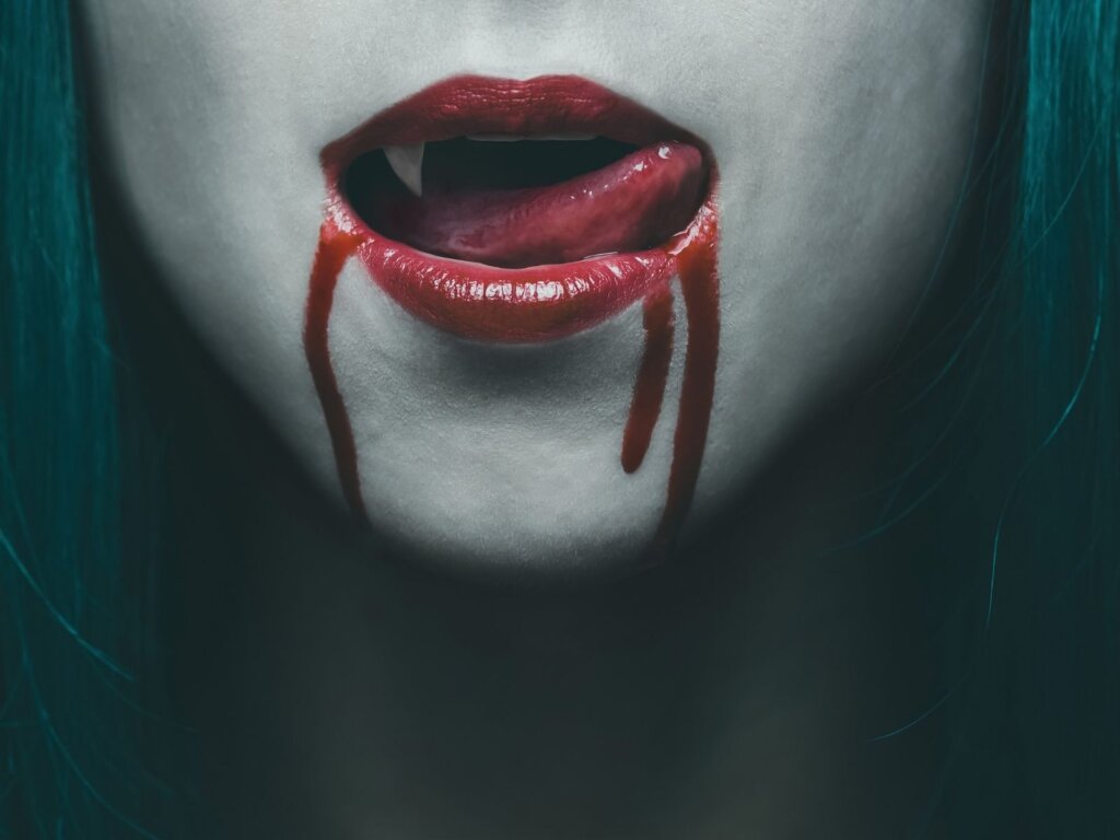 Photo of a womans mouth with sharp incisor teeth, red lips and blood dribling down chin. 