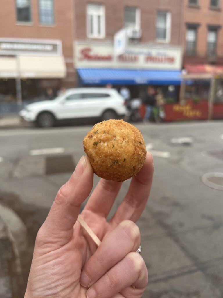 A photo of me holding up a arancini ball with Faicco's deli in the background