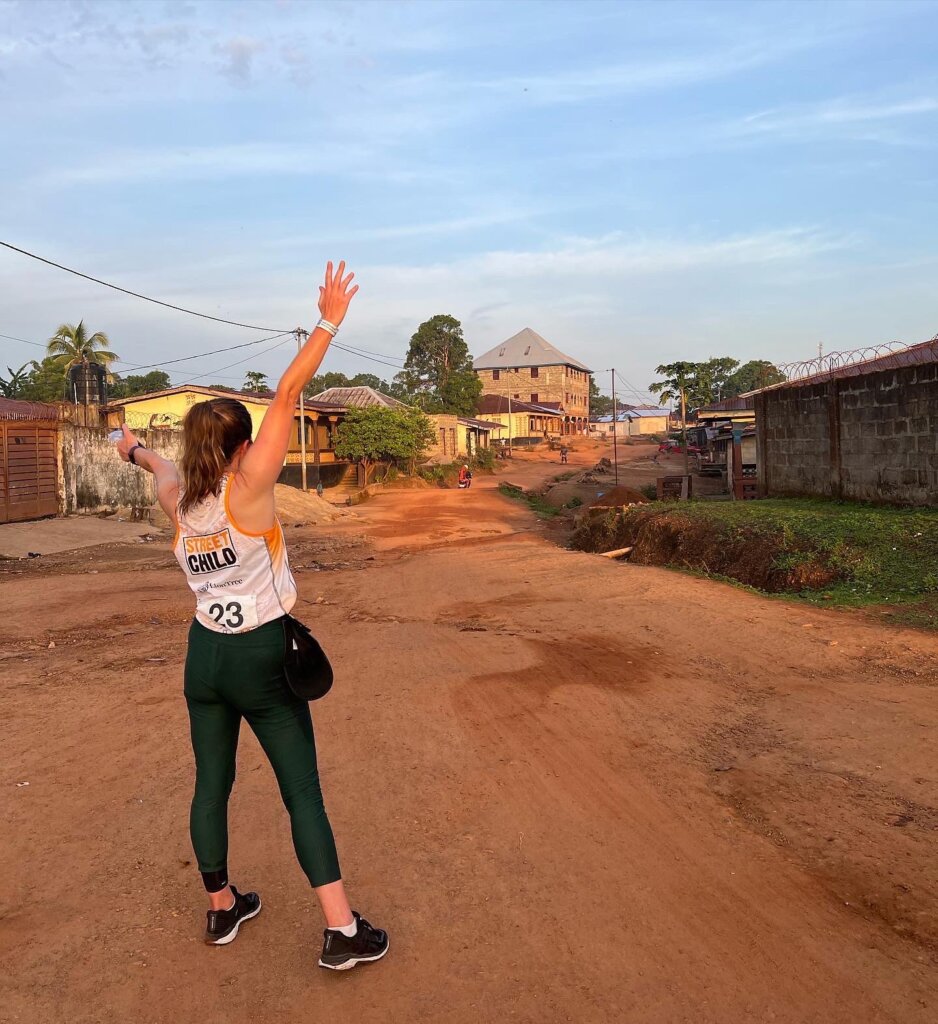 Half marathon runner Bianca with her hands in the air looking back at the red dirt street heading out of Makeni that we had just run along. 