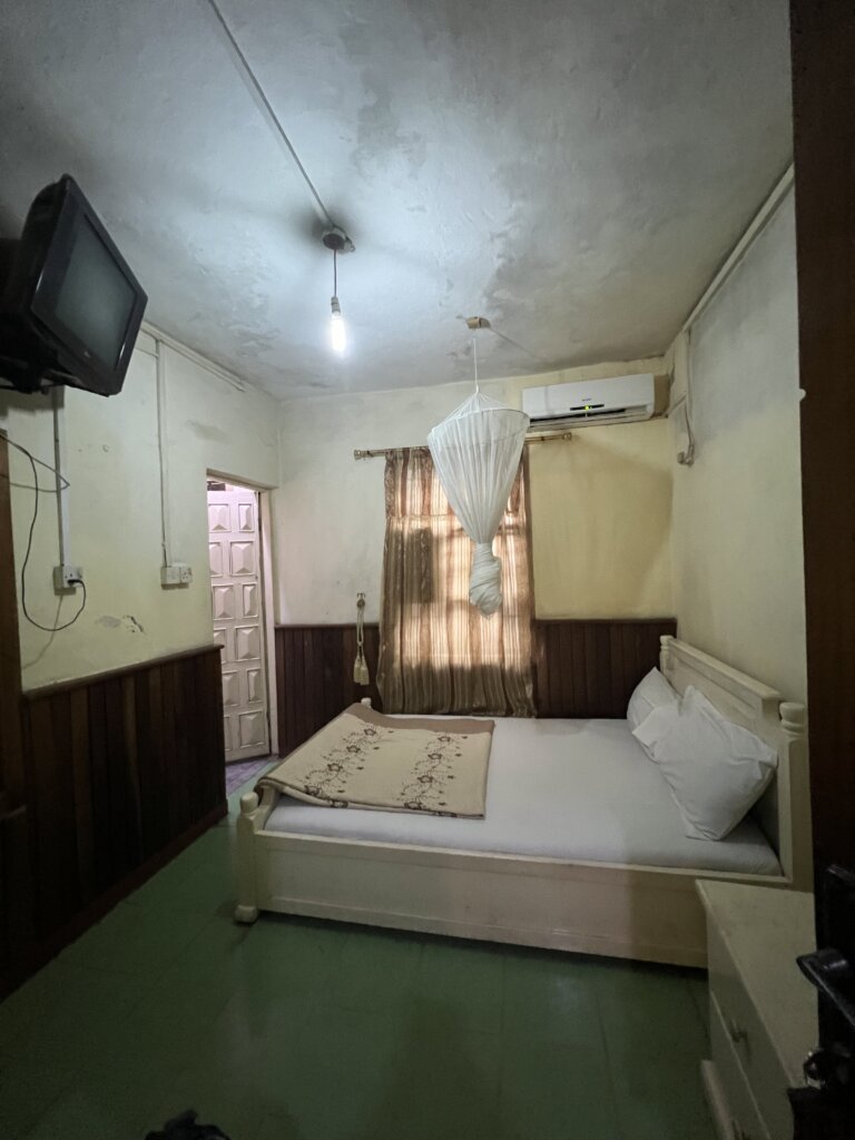 A yellow stained wall, blood splattered with squashed mosquitos. A brown curtain. A mosquito net wrapped above the bed. An old thick TV mounted on the wall. Green floor. 