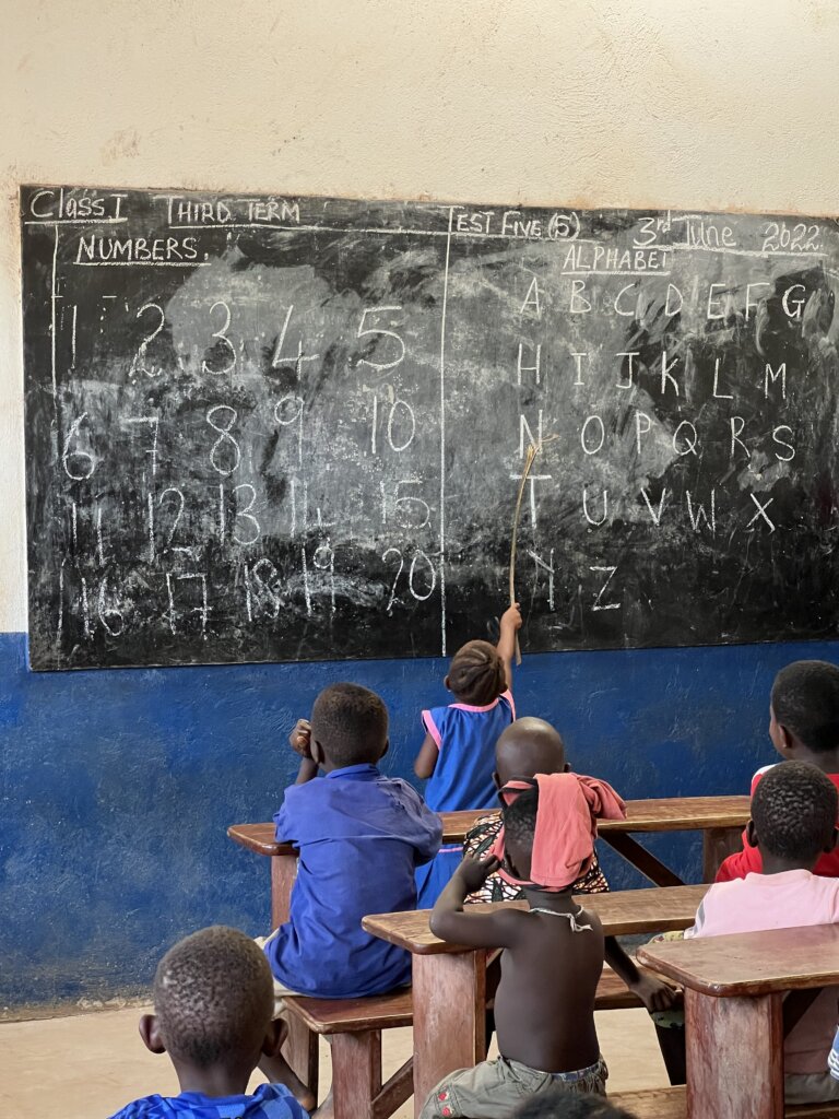 A tiny little girl stands at the blackboard spelling a word