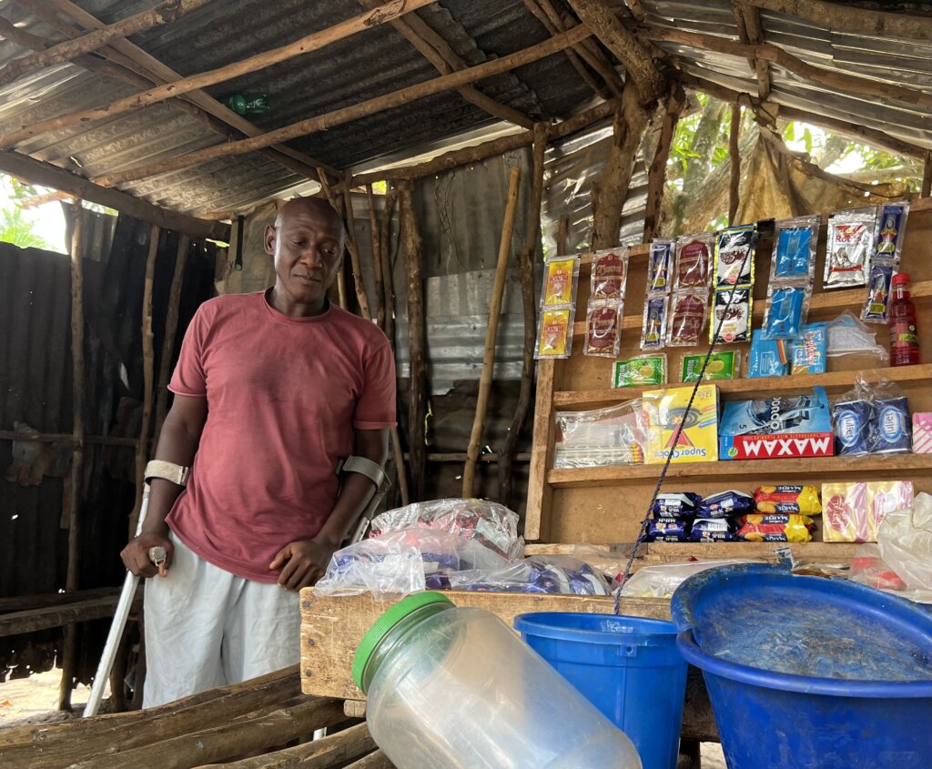 A man standing with the help of crutches surveying his little box shop. Another Street Child beneficiery glad to see the marathon runner raising funds