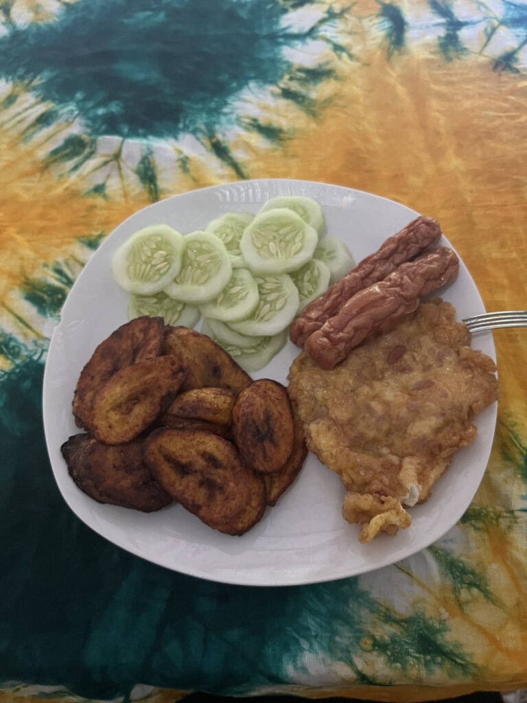 A big plate of breakfast with plantain, cucumber, two sausages and an omelette