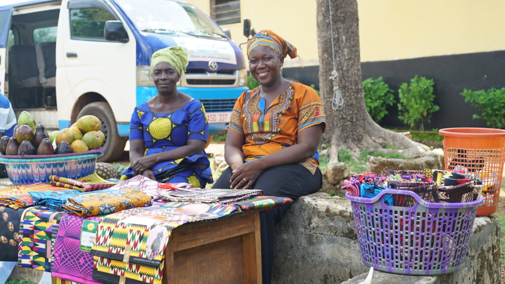 Two smiling ladies showcasing their goods for sale. There if fruit and clothing. 