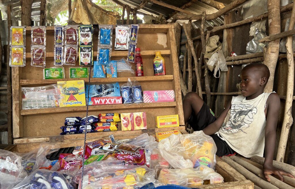 A boy sits perched next to a box that contains a selections of goods available in what is the local shop. Choice is limited. 