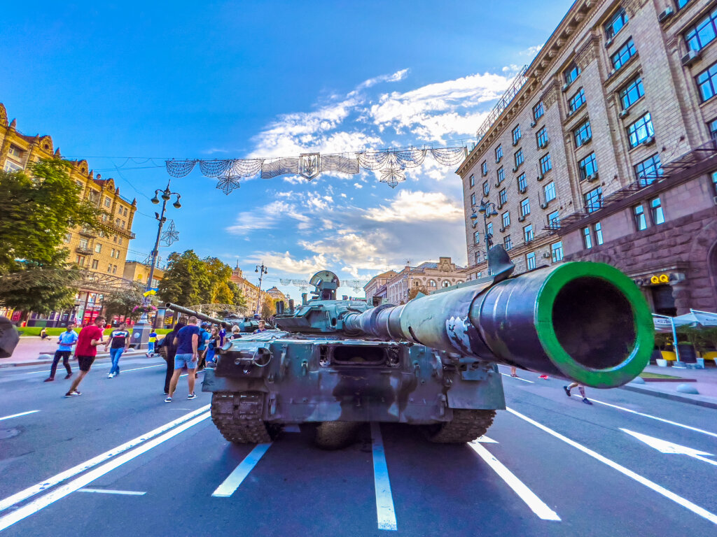 A broken Russian tank stands in the middle of the road as Ukranians walk around it as they go about their normal business