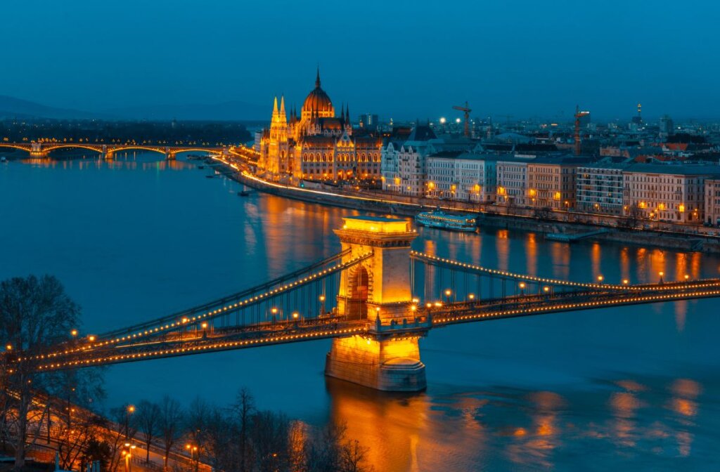 A photo of the iconic Budapest Parliament building taken from across the river with a beautifully lite up bridge  in the foreground