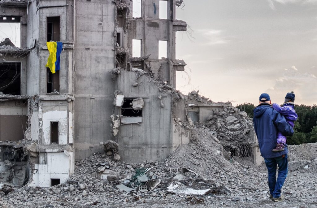 A man holding a young kid walks past a demolish building with a Ukrainian flag slumped out of a window