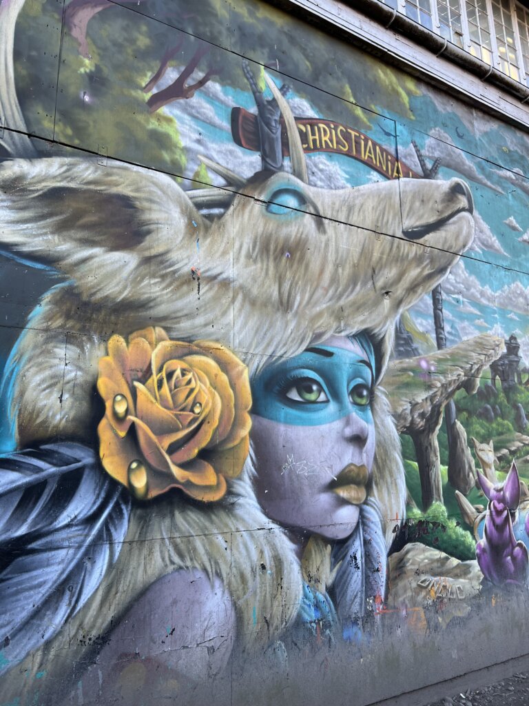 A mural of a lady wearing a dead deer as a hat with the welcome sign to Christiania painted in the background