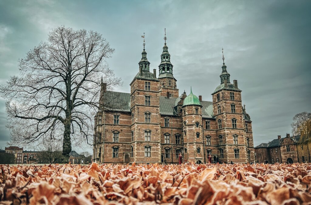 A moody shot of Rosenborg castle. A walk through the garden is free, and the views are splending. The sky is storming and the foreground appear to be leaves on the ground. A bare tree as big as the castle sit to the side.